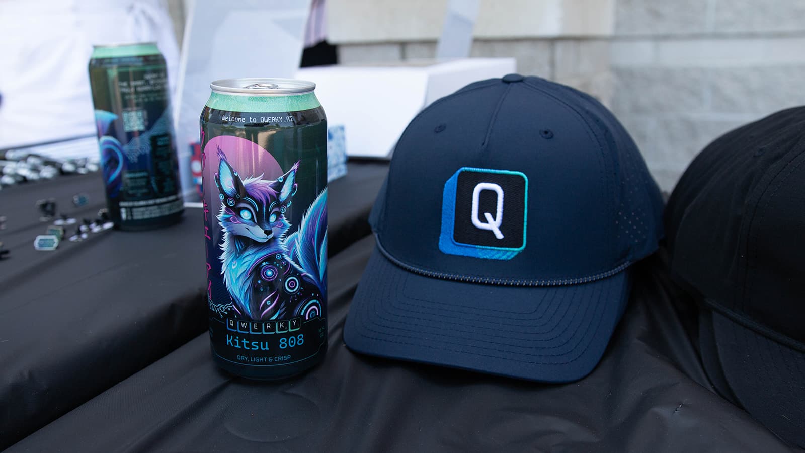 QWERKY AI single Q keyboard logo on a navy baseball hat next to a can of Kitsu 808 beer on a table of other QWERKY AI swag